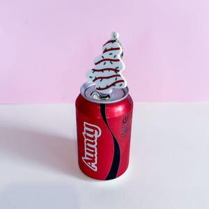 Puede incluir: Una lata de refresco roja con la palabra "Aunty" en escritura blanca. Un adorno en forma de &aacute;rbol de Navidad blanco con detalles rojos y verdes se encuentra encima de la lata abierta. El fondo es rosa y blanco.
