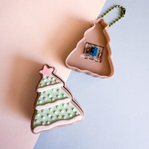 May include: Two Christmas tree-shaped keychains. One is a light green color with pink polka dots and a pink star on top. The other is a tan color with a blue switch and a green chain. The keychains are on a light pink and blue background.