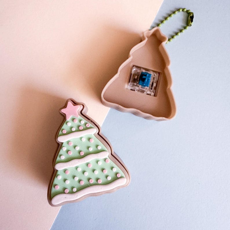 May include: Two Christmas tree-shaped keychains. One is a light green color with pink polka dots and a pink star on top. The other is a tan color with a blue switch and a green chain. The keychains are on a light pink and blue background.