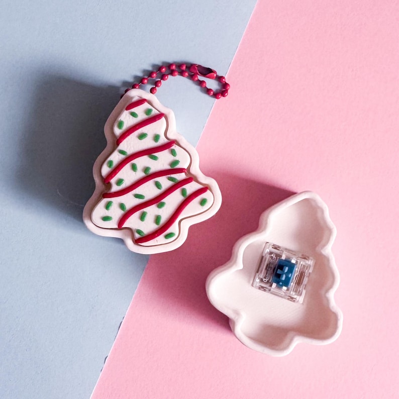 May include: A white Christmas tree-shaped keychain with red and green accents, and a matching tree-shaped container with a small blue object inside. The keychain has a red chain and is set against a pink and blue background.