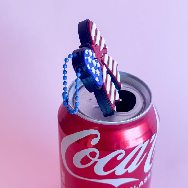 May include: A red Coca-Cola can with a decorative butterfly bottle stopper. The butterfly design features red and white stripes, blue stars, and a blue chain. The background is a solid pink.