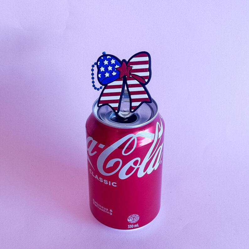 May include: A red Coca-Cola Classic can with a decorative bow featuring the American flag design. The bow is blue with white stars and red and white stripes. The can is 330 ml.