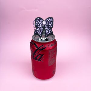Puede incluir: Una lata roja de Coca-Cola con un adorno en forma de lazo blanco y negro. El lazo tiene un dise&ntilde;o de estampado de leopardo con manchas moradas. El fondo es rosa.