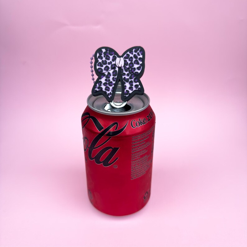 Puede incluir: Una lata roja de Coca-Cola con un adorno en forma de lazo blanco y negro. El lazo tiene un dise&ntilde;o de estampado de leopardo con manchas moradas. El fondo es rosa.