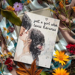 以下が含まれることがあります： 「just a girl who loves Kdramas」のテキストとキスをしているカップルのイラストが描かれた本。本は花とチェック柄の毛布に囲まれています。
