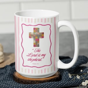 May include: White ceramic mug with a pink and white striped pattern. The mug features a framed cross design with a patchwork pattern and the text "The Lord is my shepherd." Psalm 23:1 is also printed on the mug.