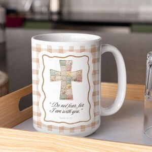 Può includere: Tazza in ceramica bianca con motivo a quadretti e disegno a croce. La croce è realizzata in tessuto patchwork di vari colori. La tazza presenta il testo "Do not fear, for I am with you." e "Isaiah 41:10".