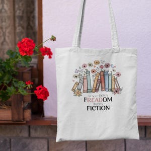 May include: Cream tote bag featuring a colourful illustration of books, flowers, and butterflies. The text on the bag reads "Where there's FREADOM there's FICTION". The bag has two white handles.