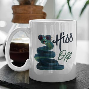 Op de afbeelding: Witte keramische mok met een cartoon slang met een zonnebril. De slang is opgerold en is groen en blauw. De mok heeft de woorden "Hiss Off" in zwarte en groene letters.