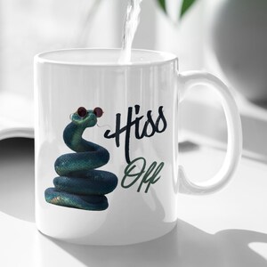Op de afbeelding: Witte keramische mok met een opgerolde blauwe slang met zonnebril. De mok heeft de tekst "Hiss Off" in zwarte en groene letters. Een straal water wordt in de mok gegoten.