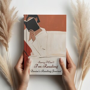 May include: A hardcover reading journal with a terracotta and cream-colored cover featuring an illustration of a person reading a book. The text on the cover reads, Sorry I Can't I'm Reading Denise's Reading Journal in dark brown script.