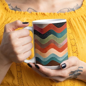 Puede incluir: Taza de cerámica blanca con un patrón ondulado colorido en tonos marrones, naranjas, verdes y azules. La taza es sostenida por una persona que lleva una blusa amarilla. La taza tiene un asa blanca.