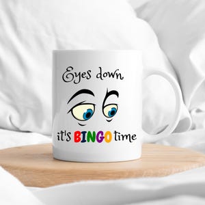 Könnte beinhalten: Weiße Keramik-Tasse mit dem Text "Eyes down, it's BINGO time" in schwarzen und farbigen Buchstaben. Die Tasse zeigt Cartoon-Augen mit blauen Iris. Die Tasse steht auf einer Holzoberfläche.