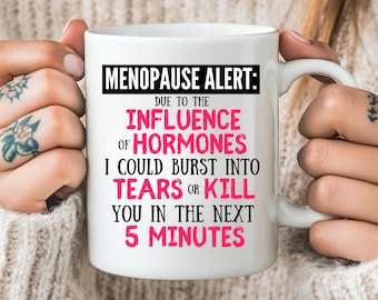 May include: White ceramic mug with the text "MENOPAUSE ALERT: DUE TO THE INFLUENCE OF HORMONES I COULD BURST INTO TEARS OR KILL YOU IN THE NEXT 5 MINUTES" in black and pink lettering. The mug is held by a person.