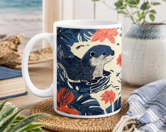 Può includere: Una tazza in ceramica bianca con un'illustrazione grafica di una lontra dal pelo scuro che nuota tra foglie tropicali blu scuro e vivaci fiori color corallo su uno sfondo color crema chiaro con un manico curvo sul lato sinistro.