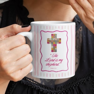 Può includere: Tazza in ceramica bianca con un motivo a righe rosa e bianche. La tazza presenta un disegno a croce incorniciata in rosa con un motivo patchwork. Il testo "The Lord is my shepherd" è stampato sotto, con "Psalm 23:1".