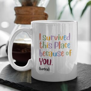 May include: White ceramic mug with the text "I survived this Place because of You" in colourful letters. The word "barley" is in parentheses below the text. The mug has a large handle and is set against a blurred background.