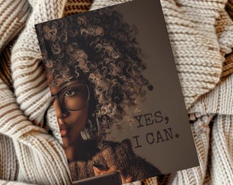 Könnte beinhalten: Ein Notizbuch mit einem Porträt einer Frau mit Brille und dem Text "YES, I CAN." Die Frau hat lockiges Haar und hält ein Telefon. Das Notizbuch liegt auf einer beigen Strickdecke.