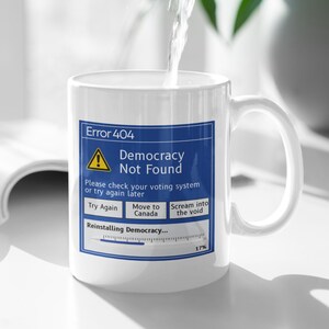May include: White ceramic mug with a blue and white graphic that reads "Error 404 Democracy Not Found." The mug has a handle and is being filled with water. The graphic includes the text "Try Again," "Move to Canada," and "Scream into the void."