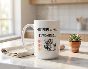 May include: A white ceramic mug featuring the black text "MENOPAUSE ALERT: THIS WOMAN IS..." followed by the word "HOT" in red, orange, and yellow, and a black and white illustration of a person sitting in front of a small electric fan.