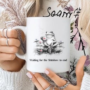 Puede incluir: Taza de cerámica blanca con una ilustración en blanco y negro de una rana sosteniendo una taza, sentada en un tronco con setas. La taza tiene el texto "Waiting for the Shitshow to end" en negro.