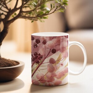 May include: White ceramic mug featuring a floral design in shades of pink, burgundy, and gold. The design includes stylized flowers and berries. The mug has a curved handle and is set against a blurred background.
