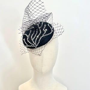 May include: A black fascinator with a decorative design of white rhinestones and a black netting veil. The headpiece is displayed on a mannequin head against a white background. The fascinator is an elegant accessory.
