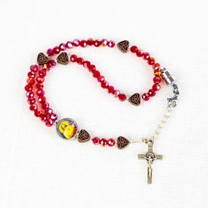Könnte beinhalten: Ein roter Rosenkranz mit einer silbernen Kreuzigung und einem kleinen religiösen Bild. Die Perlen sind tiefrot, mit braunen dekorativen Elementen. Der Rosenkranz befindet sich auf weißem Hintergrund.