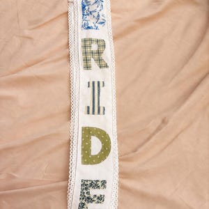 May include: A long white fabric sash featuring the word BRIDE appliqued in vertical order. Each letter is crafted from a different patterned fabric, including blue floral, green plaid, blue striped, olive polka dot, and dark green leaf prints.
