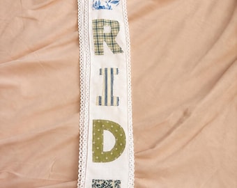 May include: A long white fabric sash featuring the word BRIDE appliqued in vertical order. Each letter is crafted from a different patterned fabric, including blue floral, green plaid, blue striped, olive polka dot, and dark green leaf prints.