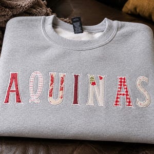 May include: A light gray crewneck sweatshirt featuring the word AQUINAS appliqued across the chest in various fabric patterns including red floral, pink stripes, red gingham, and cream with small red flower accents, with a black neck label.