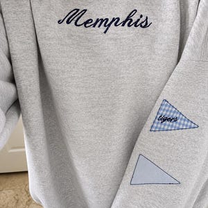 Op de afbeelding: Een lichtgrijs sweatshirt met ronde hals en het woord Memphis in donkerblauw cursief geborduurd op de borst. De mouw is voorzien van twee driehoekige patches, één met een blauw-wit ruitpatroon met de tekst tigers en één effen lichtblauw.