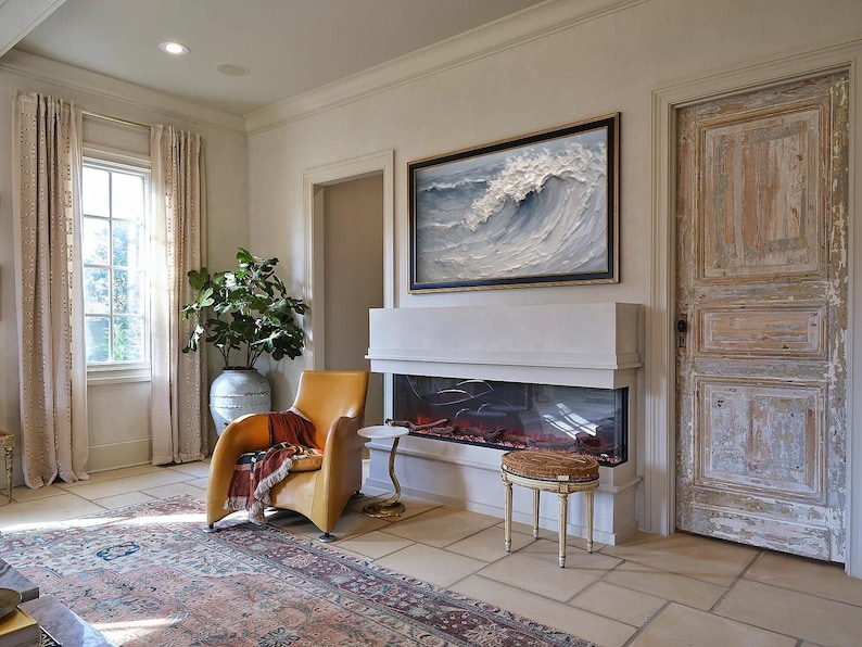 Op de afbeelding: Een woonkamer met een moderne open haard, een gele leren fauteuil en een groot ingelijst schilderij van een golf. De kamer heeft een vloerkleed, een bijzettafel, een decoratieve kruk en een grote plant in een keramische vaas. De deur is verweerd.