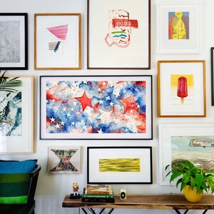 May include: A gallery wall with a variety of framed artwork, including abstract paintings, a watercolor painting of stars and stripes, and a framed print of a popsicle. The wall is painted white and there is a wooden table with books and a plant on it.