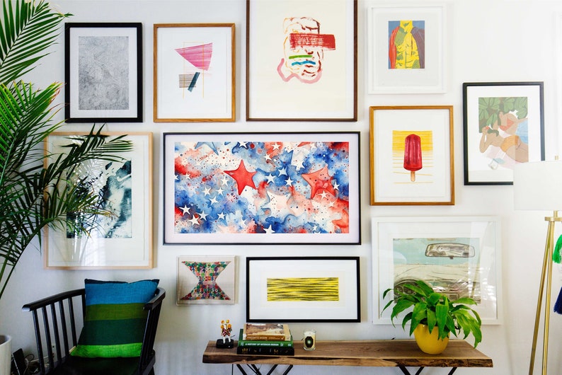 May include: A gallery wall with a variety of framed artwork, including abstract paintings, a watercolor painting of stars and stripes, and a framed print of a popsicle. The wall is painted white and there is a wooden table with books and a plant on it.