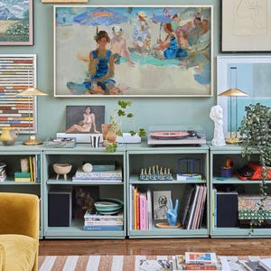Op de afbeelding: Een woonkamer met lichtgroene opbergmeubels en een bijpassende muur. De kamer heeft een groot ingelijst schilderij van een strandsc&egrave;ne, een platenspeler, boeken en decoratieve items. Een gele stoel en een gestreept tapijt voegen kleur toe.