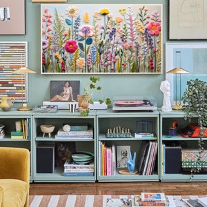 May include: A light blue bookshelf with three shelves, filled with books, plants, and decorative items. The bookshelf is against a light blue wall with a framed floral embroidery hanging above it.