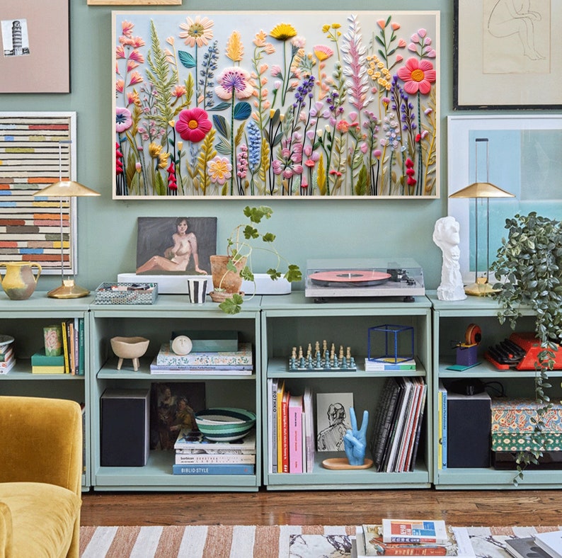 May include: A light blue bookshelf with three shelves, filled with books, plants, and decorative items. The bookshelf is against a light blue wall with a framed floral embroidery hanging above it.