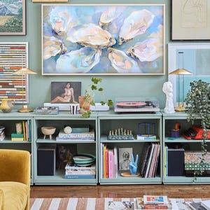 Op de afbeelding: Een abstract schilderij van oesters in een lichtblauw en wit kleurenpalet hangt boven een mintgroene opbergkast. De kast bevat boeken, een platenspeler en decoratieve items. Een gouden lamp en een beeldhouwwerk zijn ook zichtbaar.