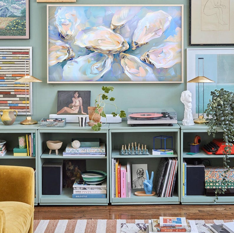 Op de afbeelding: Een abstract schilderij van oesters in een lichtblauw en wit kleurenpalet hangt boven een mintgroene opbergkast. De kast bevat boeken, een platenspeler en decoratieve items. Een gouden lamp en een beeldhouwwerk zijn ook zichtbaar.