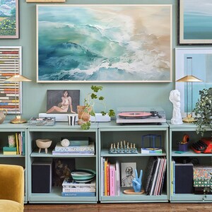 May include: A set of four teal blue bookshelves with various items on the shelves, including books, records, a record player, a plant, and decorative items. The shelves are against a light blue wall with artwork above them.