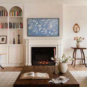 Op de afbeelding: Woonkamer met open haard, boekenkasten en salontafel. Boven de open haard hangt een blauw schilderij van witte vogels in vlucht. Op de salontafel staan een brandende kaars en een vaas met bloemen.