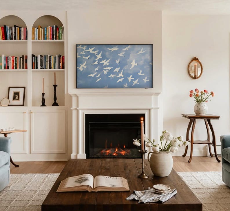 Op de afbeelding: Woonkamer met open haard, boekenkasten en salontafel. Boven de open haard hangt een blauw schilderij van witte vogels in vlucht. Op de salontafel staan een brandende kaars en een vaas met bloemen.