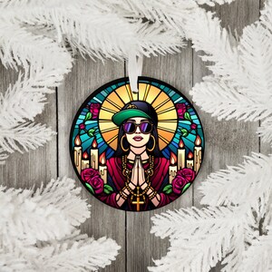 May include: Round Christmas bauble depicting a stained glass style illustration of a woman wearing a green baseball cap, sunglasses, and a gold chain. She is holding her hands in a prayer position and is surrounded by candles and roses.