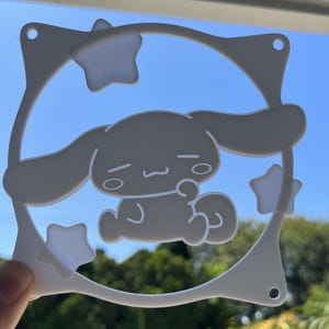 May include: A white, die-cut decorative piece featuring a cartoon character within a circular frame adorned with stars. The character has large ears and a relaxed expression. The piece has three small holes.