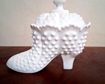 May include: A white, hobnail-textured glass shoe-shaped trinket box with a matching lid. The lid has a rounded finial. The shoe has a high heel and is sitting on a dark wooden surface. The overall design is vintage.