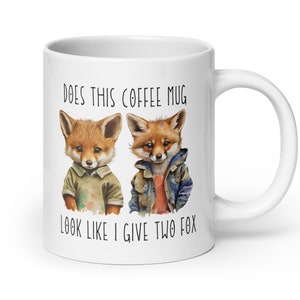 Könnte beinhalten: Weiße Keramik-Kaffeetasse mit zwei Cartoon-Füchsen in Kleidung. Der Text auf der Tasse lautet: "DOES THIS COFFEE MUG LOOK LIKE I GIVE TWO FOX".