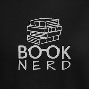 May include: Black sweatshirt with white embroidered design. The design features a stack of books above the words "BOOK NERD" in a sans-serif font. The "OO" in "BOOK" is replaced with glasses.
