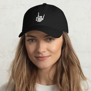 Könnte beinhalten: Schwarze Baseballkappe mit einer weißen gestickten Skeletthand, die das Rock'n'Roll-Zeichen macht.