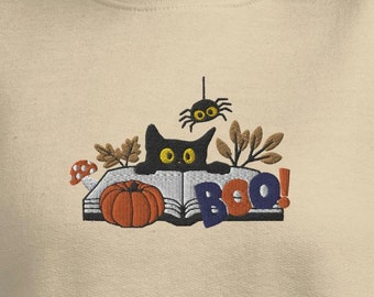 May include: A beige sweatshirt with an embroidered design of a black cat peeking over a book with a pumpkin and a spider. The text "BOO!" is embroidered on the book.